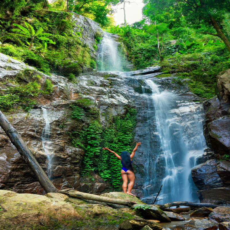 Young Female Tourist Standing Before A Waterfall Holding Her Arms Up, Costa Rica, Central America