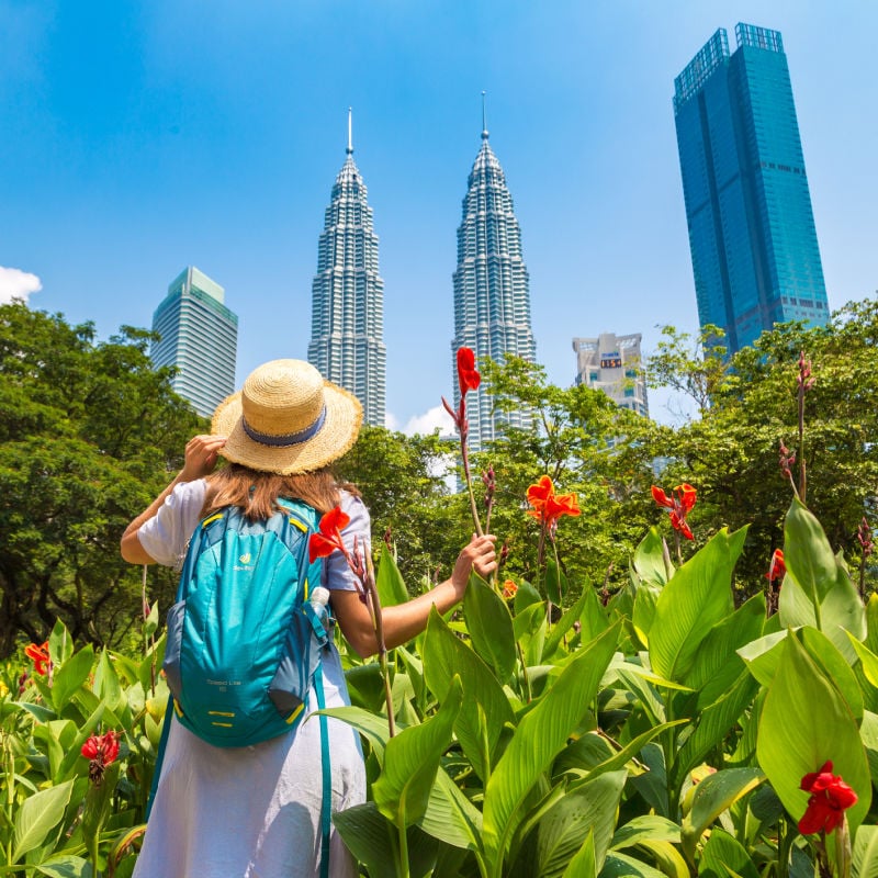 Female Backpack Walking Through A Garden With The Petronas Towers For Background, Kuala Lumpur, Malaysia, Southeast Asia